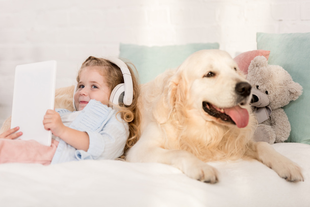 schattig kind luisteren van muziek met tablet en leunend op gouden retriever op bed in de kinderkamer - Foto, afbeelding