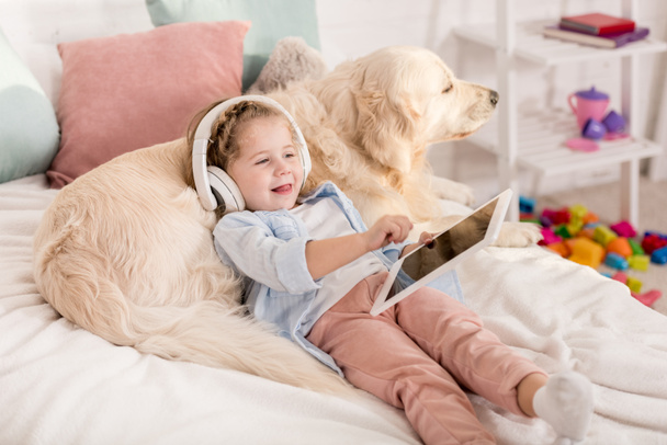 adorable cheerful kid listening music with tablet and leaning on golden retriever on bed in children room - Photo, Image