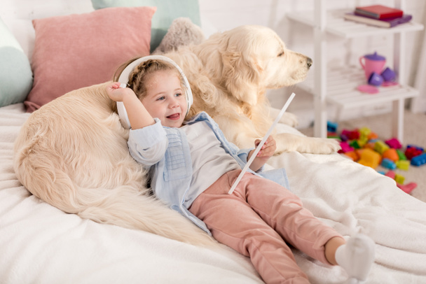 adorable kid using tablet and leaning on golden retriever on bed in children room - Foto, imagen
