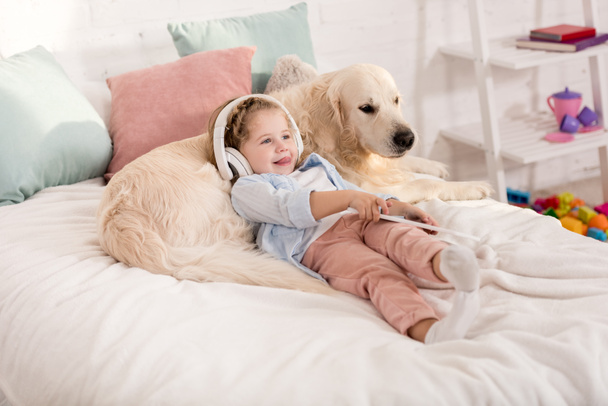 adorable kid listening music with tablet and leaning on fluffy golden retriever on bed in children room - Photo, Image