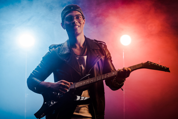 riso jovem músico masculino em jaqueta de couro realizando na guitarra elétrica no palco com fumaça e iluminação dramática
  - Foto, Imagem