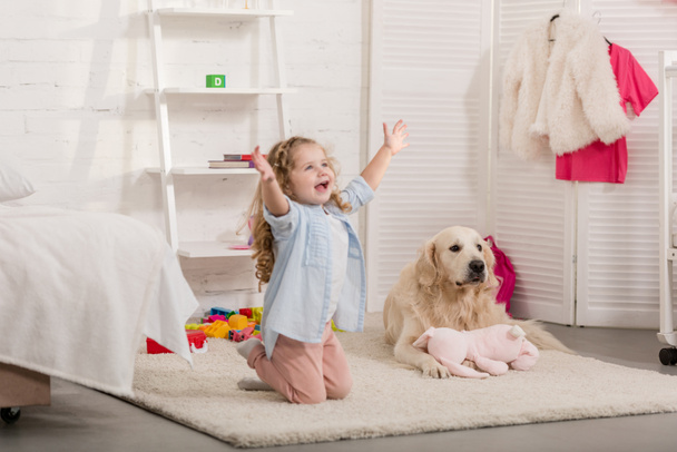 glückliches entzückendes Kind, das mit offenen Armen auf Knien steht, Golden Retriever auf Teppich im Kinderzimmer liegend - Foto, Bild