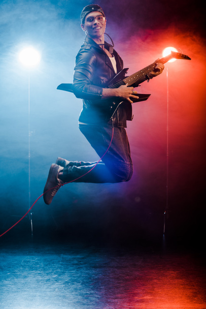 happy male musician jumping and performing on electric guitar during rock concert on stage with smoke and spotlights  - Zdjęcie, obraz