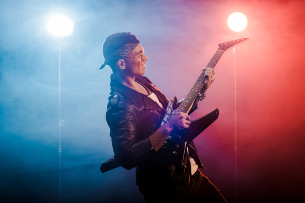 happy male rock star in leather jacket performing on electric guitar on stage with smoke and dramatic lighting  - Photo, Image