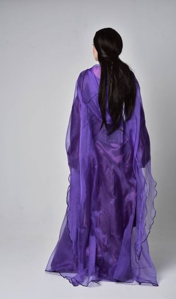 retrato de comprimento total da menina bonita com cabelo preto longo, vestindo vestido medieval fantasia roxa e capa. em pé pose no fundo do estúdio cinza
. - Foto, Imagem