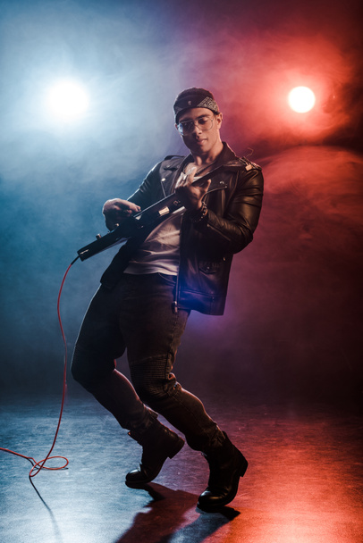 guapo rockero elegante en chaqueta de cuero que actúa en la guitarra eléctrica en el escenario con humo e iluminación dramática
  - Foto, Imagen