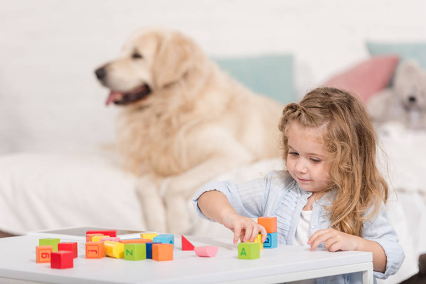 selective focus of adorable kid playing with educational cubes, golden retriever lying on bed in children room - Fotó, kép