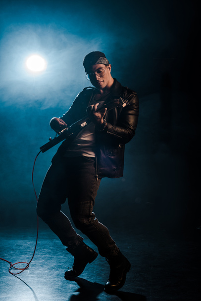 concentrated male rock star in leather jacket performing on electric guitar on stage with smoke and spotlight - Foto, afbeelding