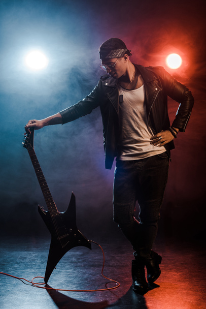 young mixed race rock musician in leather jacket posing with electric guitar on stage with smoke and dramatic lighting - Photo, Image