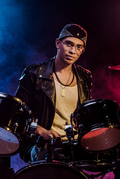 handsome mixed race male musician sitting behind drum set on stage with smoke and dramatic lighting - Photo, Image