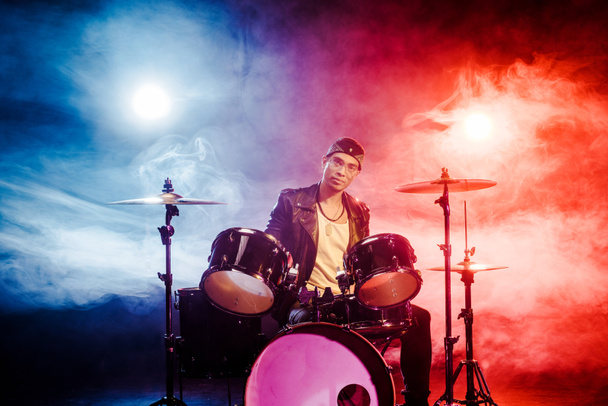 músico masculino de raza mixta seguro sentado detrás del tambor montado en el escenario con focos
 - Foto, imagen