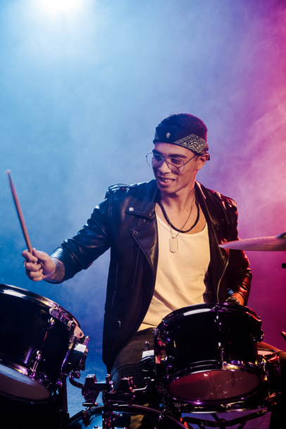 alegre joven músico masculino en chaqueta de cuero tocando la batería durante el concierto de rock en el escenario con humo e iluminación dramática
 - Foto, Imagen