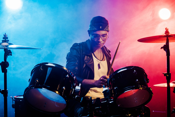 joven músico masculino enfocado en chaqueta de cuero tocando la batería durante el concierto de rock en el escenario con humo y focos
 - Foto, Imagen
