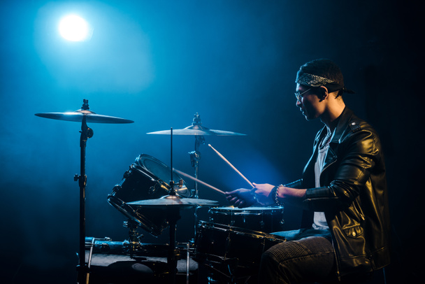 vista lateral músico masculino em casaco de couro tocando bateria durante concerto de rock no palco com fumaça e holofotes
 - Foto, Imagem