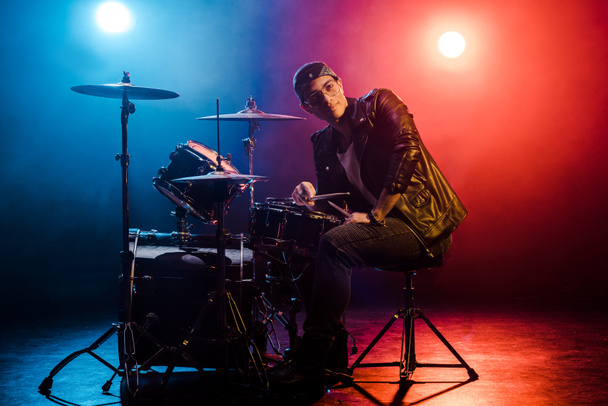 mladí Smíšené rasy mužské hudebník sedí za bicí soupravu na jevišti s reflektory a kouř - Fotografie, Obrázek