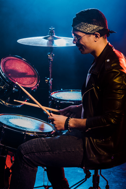 ευτυχισμένος αρσενικό μουσικός σε δερμάτινο μπουφάν παίζει ντραμς κατά τη διάρκεια συναυλίας ροκ στη σκηνή με τον καπνό και δραματικό φωτισμό  - Φωτογραφία, εικόνα