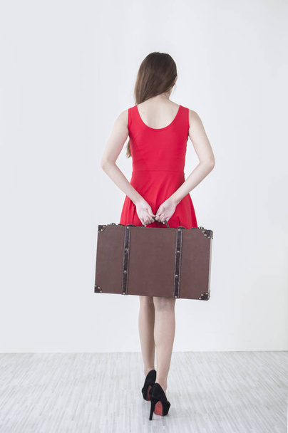 Young woman wearing a red dress holding a suit case and showing her back to the camera - Foto, Imagem
