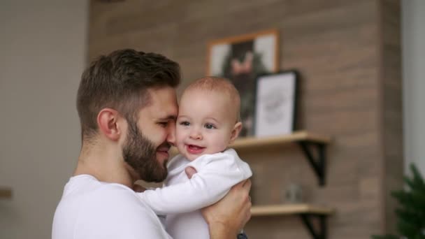 Liebender Vater im weißen T-Shirt umarmt das Baby und küsst es lachend und lächelnd auf dem Hintergrund des Weihnachtsbaums und der Girlanden - Filmmaterial, Video