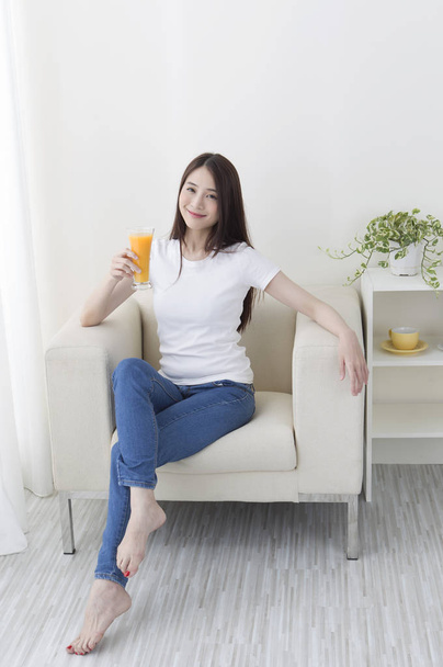 Young Asian woman in a jeans sitting on the couch holding an orange juice and smiling at the camera - Zdjęcie, obraz
