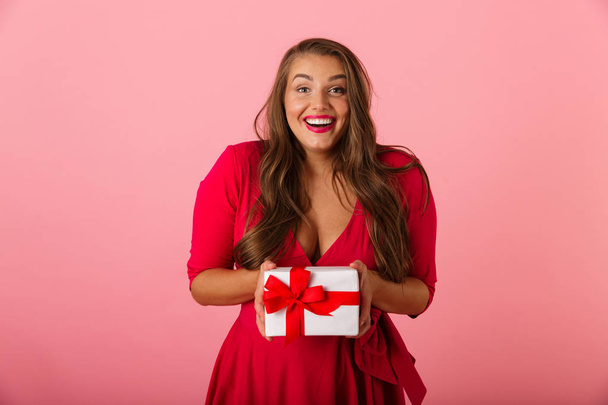 Photo of caucasian chubby woman 20s wearing red dress smiling and holding gift box isolated over pink background - Photo, Image
