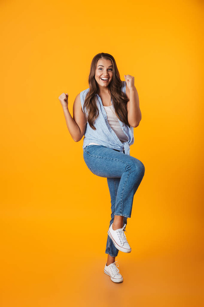 Full length portrait of an excited young casual woman standing isolated over yellow background, celebrating success - Foto, imagen