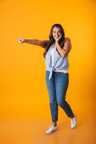 Portrait of a cheerful young casual woman standing isolated over yellow background, pointing fingers away at copy space - Foto, Imagem
