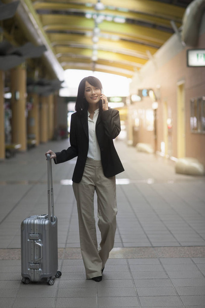 Young Asian woman wearing a suit holding a suitcase and smiling looking away - Fotó, kép