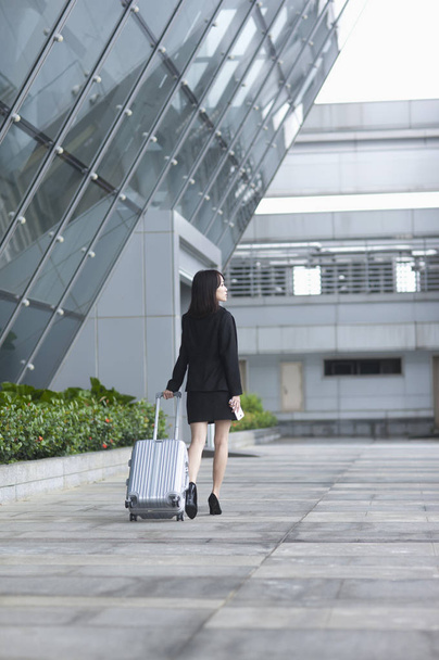 Young Asian woman in a suit holding a luggage and smiling looking away - Fotoğraf, Görsel