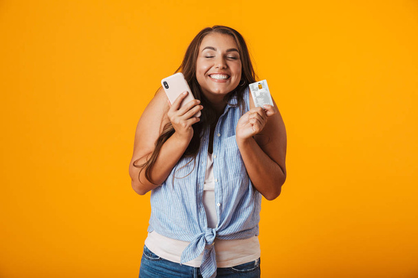 Portrait of a delighted young casual woman standing isolated over yellow background, showing plastic credit card and mobile phone - Φωτογραφία, εικόνα
