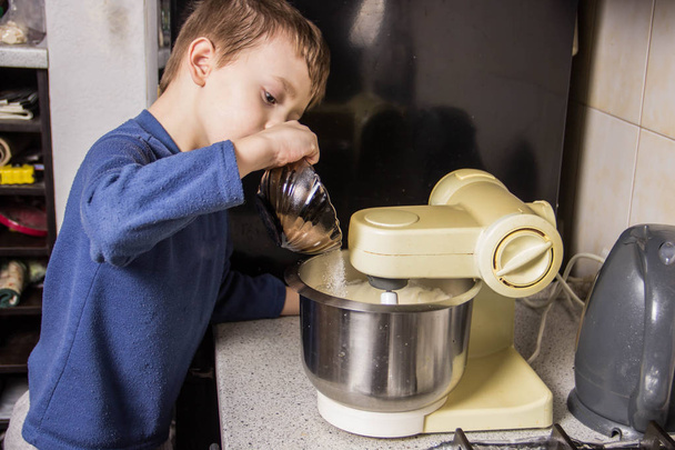 Le garçon dans la cuisine pétrit la pâte pour les cupcakes dans le mélangeur, en ajoutant des ingrédients
 - Photo, image