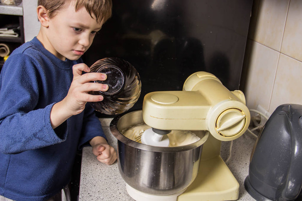 Chłopiec w kuchni wyrabia ciasto na babeczki w mikserze, Dodawanie składników - Zdjęcie, obraz