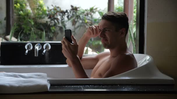 Przygnębiony nieszczęśliwy młody człowiek dostał złe wiadomości z złe wiadomości na smartfonie leżąc w łazience - Materiał filmowy, wideo