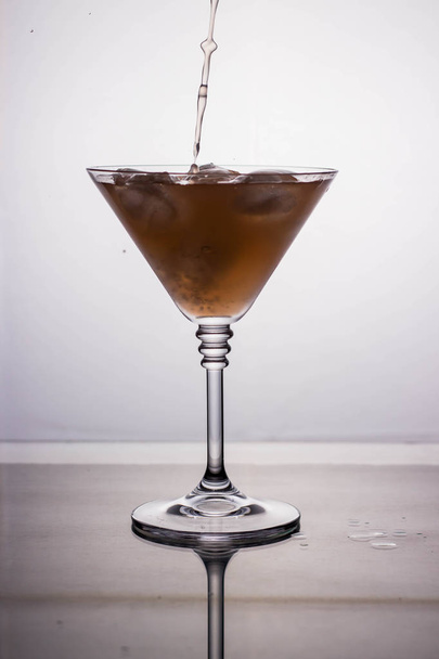 Flüssigkeit wird in ein Cocktailglas auf hellem Hintergrund mit Reflexion auf einem Spiegeltisch gegossen - Foto, Bild
