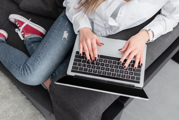 cropped view of girl sitting on couch and using laptop in living room - Foto, Imagem