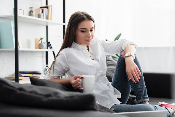 pensive girl sitting on couch and holding cup of coffee in living room - Photo, image