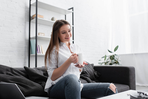 smiling girl sitting on couch and holding cup of coffee in living room - Foto, immagini