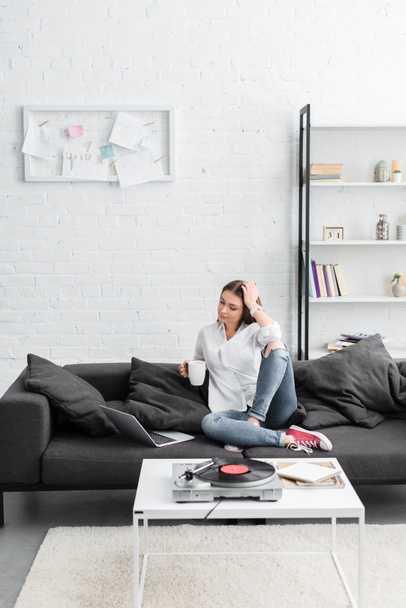 girl sitting on couch with coffee cup, using laptop and listening to vinyl record player in living room - Photo, Image