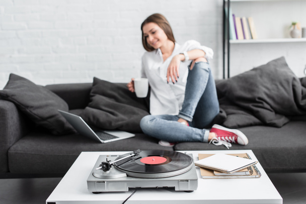 girl in white shirt sitting on couch with coffee cup, using laptop and listening to vinyl record player in living room - Zdjęcie, obraz