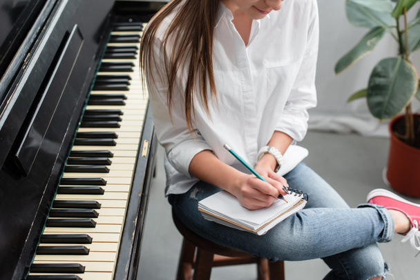 Ausgeschnittene Ansicht eines Mädchens mit Notizbuch, das neben dem Klavier sitzt und im Wohnzimmer Musik komponiert - Foto, Bild