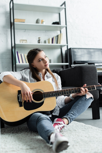 beautiful pensive girl sitting and playing guitar in living room - Fotografie, Obrázek