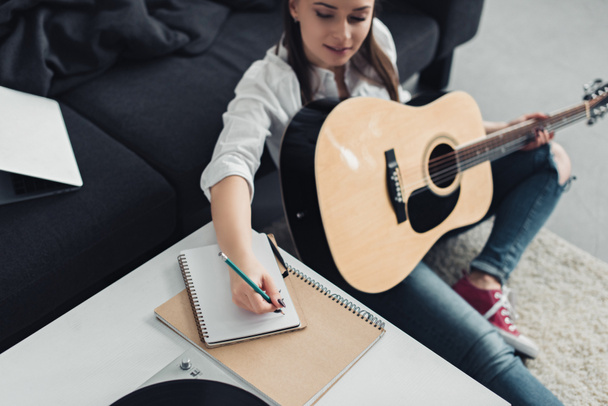girl with acoustic guitar sitting on floor and writing in notebook while composing music at home - Foto, Imagem