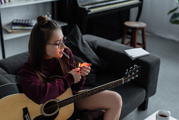 mooi meisje zitten, verlichting marihuanaverbinding aanstak en houden van gitaar thuis - Foto, afbeelding