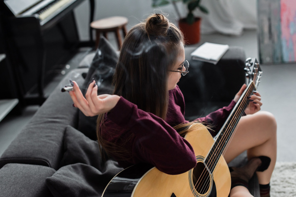 girl sitting, holding marijuana joint and playing guitar in living room - Foto, afbeelding