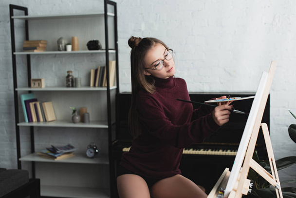 hermosa chica en jersey de color burdeos sentado y la pintura sobre lienzo en casa
  - Foto, imagen