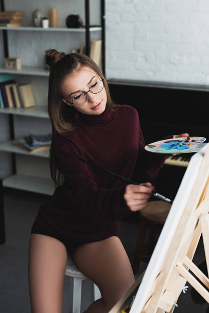 beautiful girl in burgundy sweater and glasses sitting, holding palette and painting at home  - 写真・画像