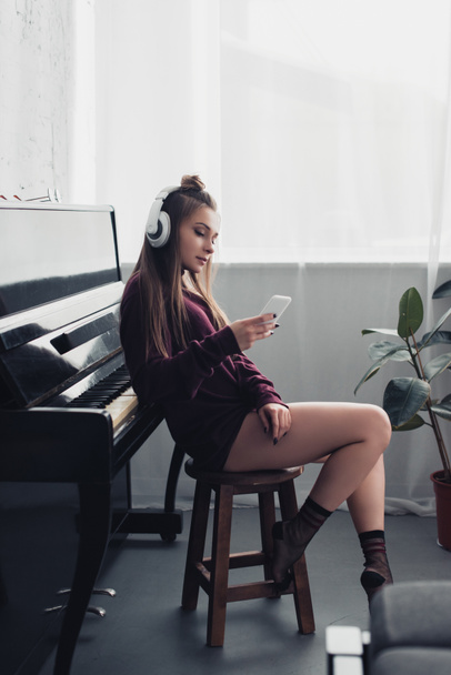 girl in headphones sitting and using smartphone in living room with piano on background - Φωτογραφία, εικόνα