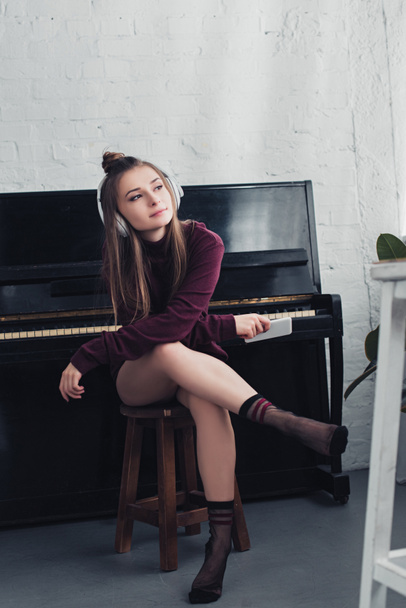 beautiful girl with headphones on head sitting on chair in front of piano and holding smartphone in living room - Foto, afbeelding