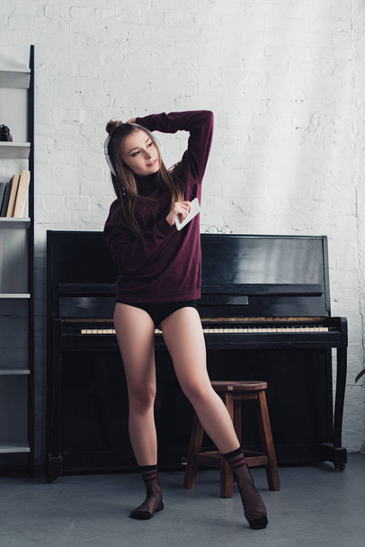 beautiful girl with headphones on head holding smartphone and standing near piano at home - Foto, afbeelding