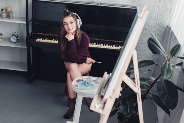attractive girl with headphones on head sitting on chair in front of piano, holding smartphone and looking at easel in living room - Photo, Image