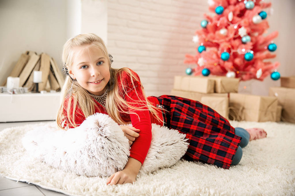 A little blonde girl in a red sweater and checkered womb sits in a bright room on the floor against the backdrop of a pink Christmas tree and hugs a large white soft pillow and looks into the camera while smiling. Christmas theme - Photo, Image